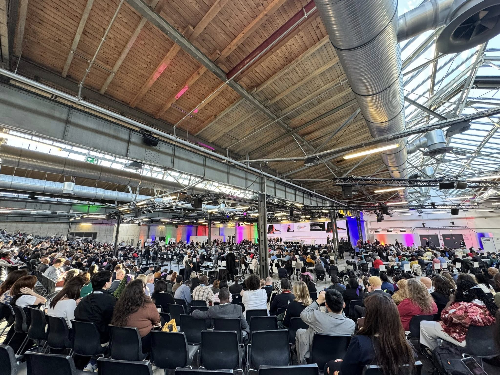A wide-angle view of the Global Disability Summit 2025 shows a large, well-attended conference hall filled with diverse participants seated and facing a central stage. The venue features high ceilings with exposed beams and industrial ductwork, and the stage area is lit with colorful lights and large screens displaying the event. The crowd includes individuals from various backgrounds, highlighting the inclusive and international nature of the summit.