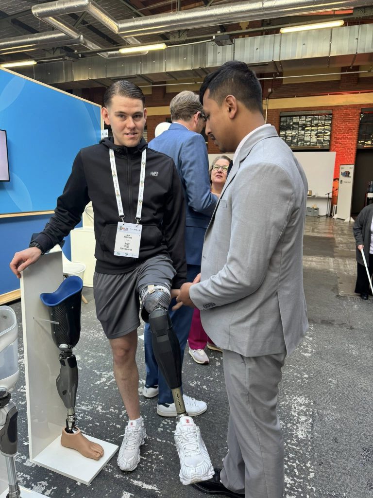 At the Global Disability Summit, DIrector of BYAN, Nir, in a light gray suit gently touches and learns about a high-tech prosthetic leg worn by another participant. The man wearing the prosthetic is dressed in a black jacket and shorts and stands next to a display featuring an additional prosthetic limb. The interaction takes place in a modern indoor venue with other attendees and exhibition booths visible in the background.