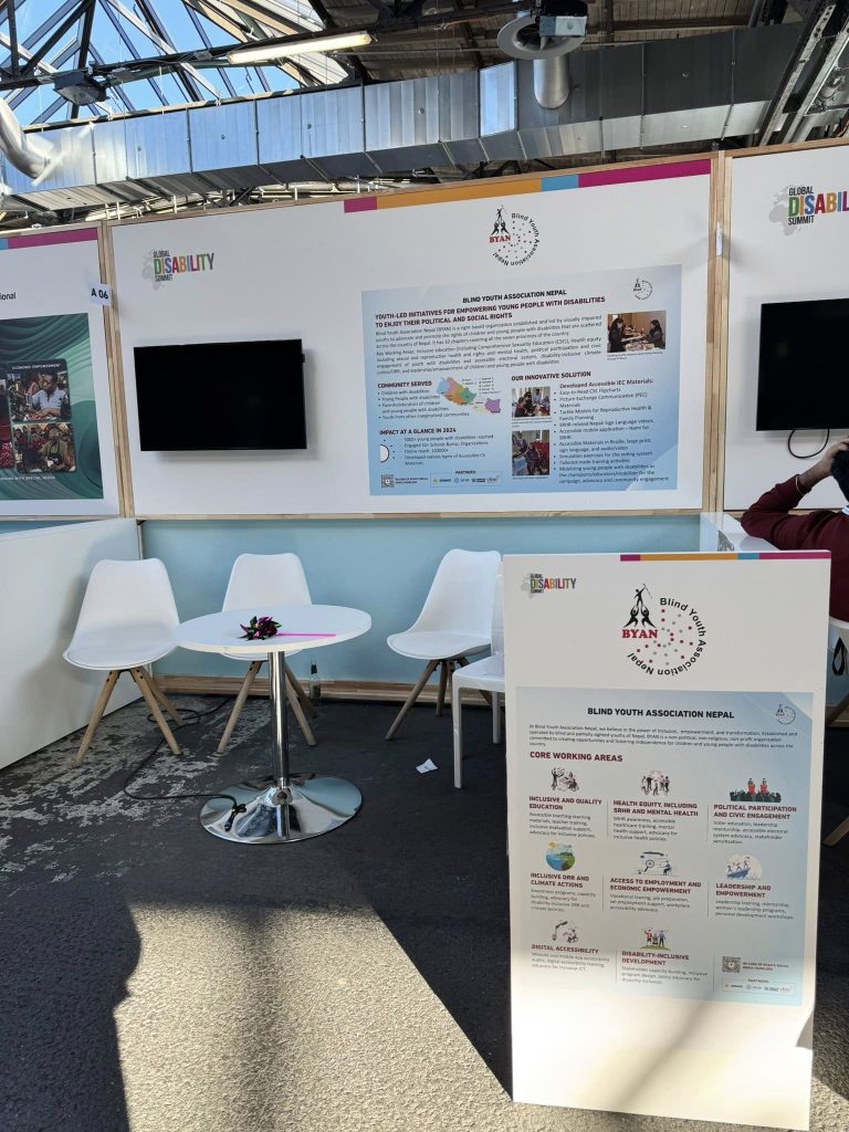 BYAN's booth at the Global Disability Summit. The display features posters detailing youth-led initiatives for empowering people with disabilities in Nepal, along with their core working areas such as inclusive education, health, civic engagement, and economic empowerment. In front of the booth are white chairs and a small table with a flower decoration and a pen on it, creating a welcoming space for discussions. There is a TV hanging on a wall along with a banner of BYAN's innovations on the side.