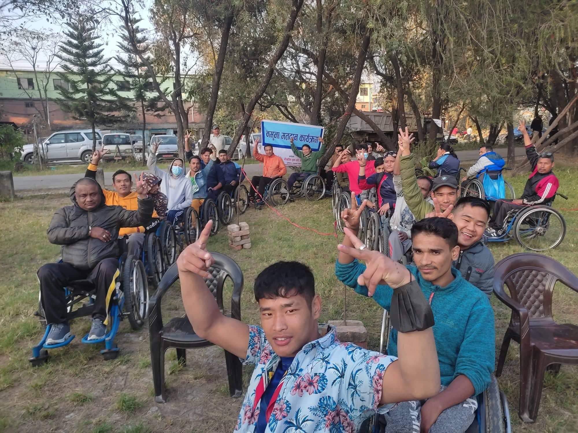 Around 20 people are seated outdoors on a grassy area under trees. Many of them are using wheelchairs. They are arranged in rows facing the camera. Several people have one or both hands raised with two fingers extended. A banner with Nepali text is placed behind the group. Plastic chairs are visible among the wheelchairs. Buildings, parked vehicles, and trees can be seen in the background.