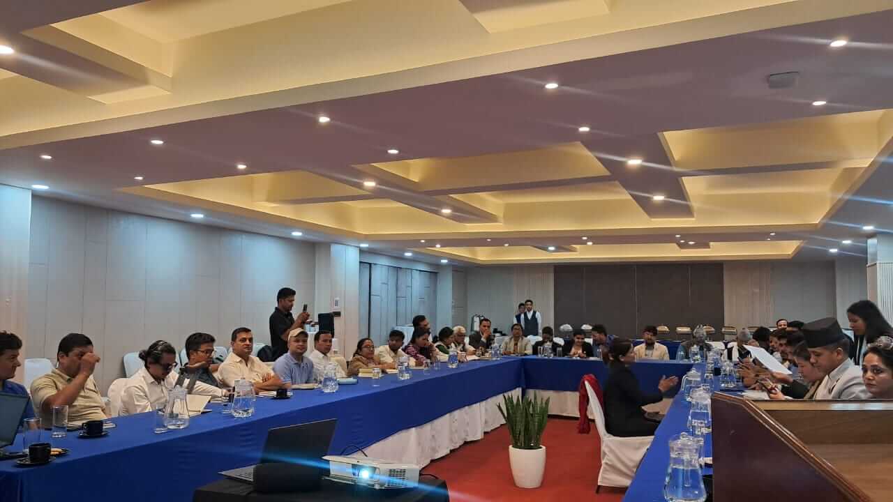 A large conference room arranged in a U-shaped seating layout with long tables covered in blue tablecloths. Around 30 participants are seated, listening to a discussion. At the front right, one participant is reading from a document while another person seated in the center appears to be speaking or interpreting. Several attendees have laptops, notebooks, water bottles, and cups on the tables. A projector is set up at the front of the room. The ceiling features recessed geometric lighting, creating a bright, professional meeting environment. A staff member stands at the back of the room recording the session on a mobile phone.
